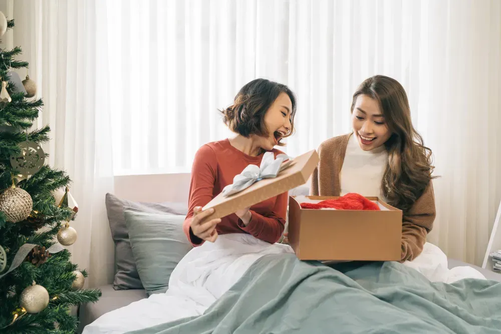 Best friends exchanging gifts during the holiday season while cozy under a blanket, on the couch. 
