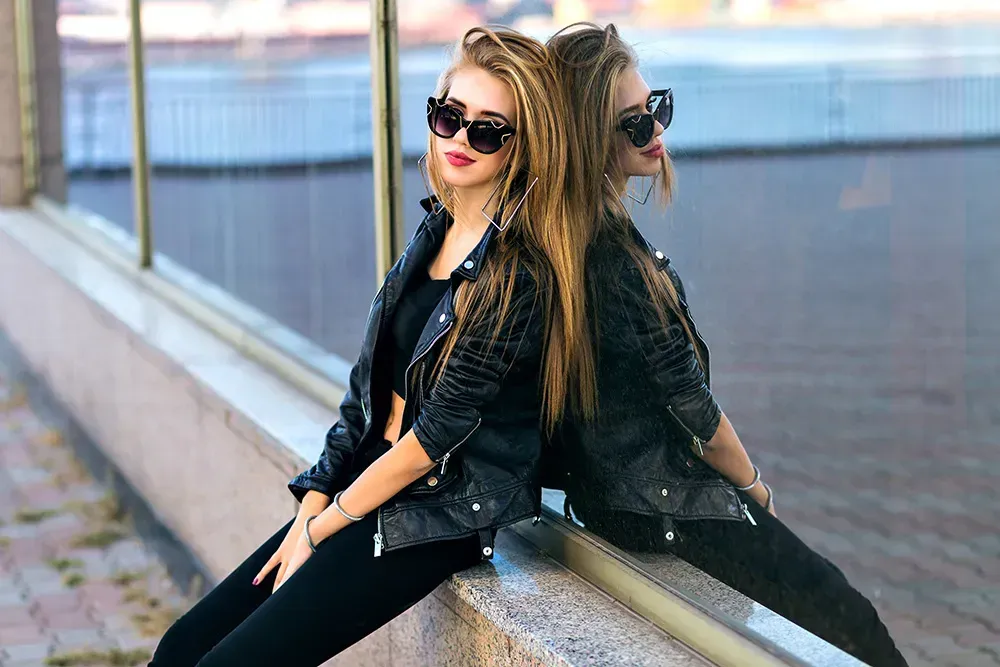 Woman wearing black leather jacket leaning against window with sunglasses on