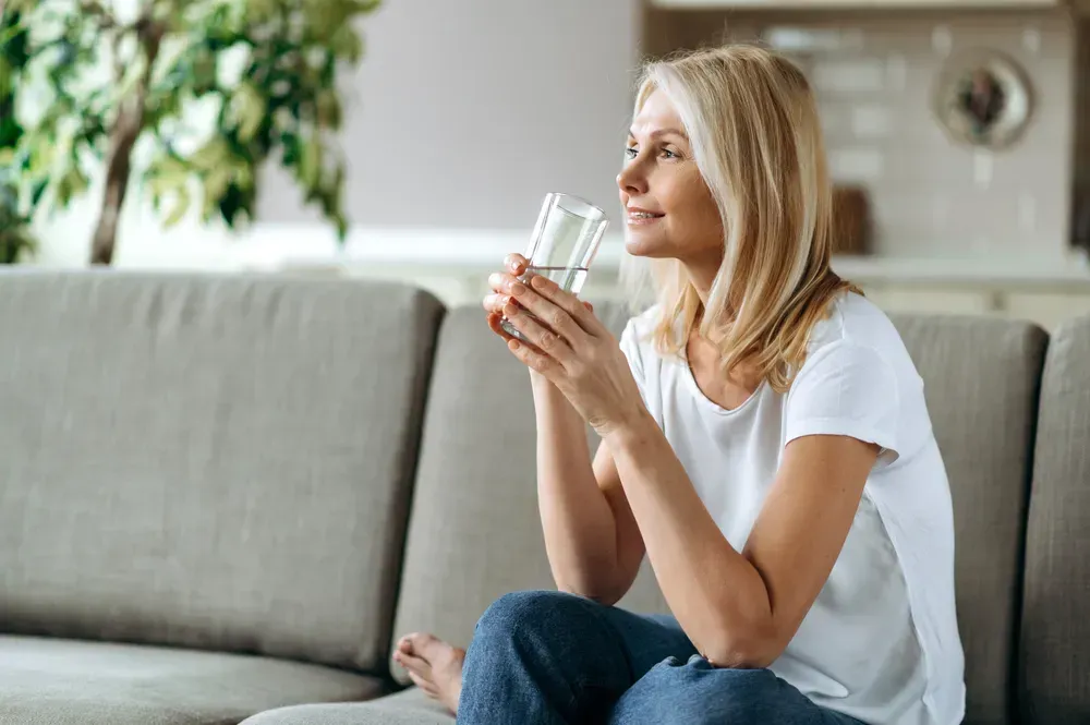 Mature white blonde dressed in casual wear sits on the couch in a living room, holding a glass of pure water in a hand and smiling.