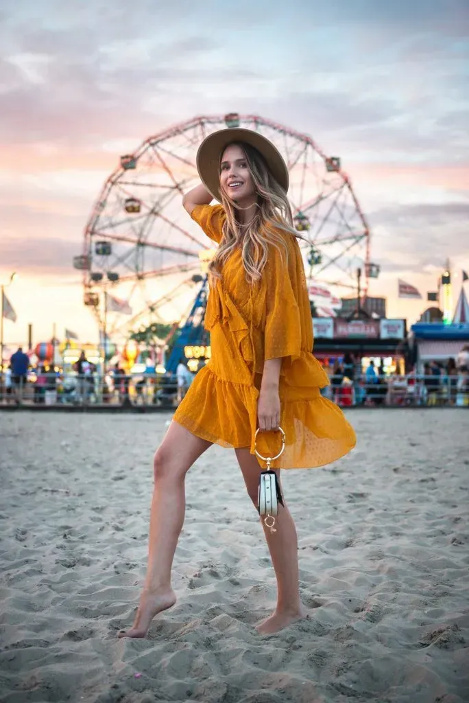 Woman in yellow dress and wide brim hat exploring carnival on the beach.