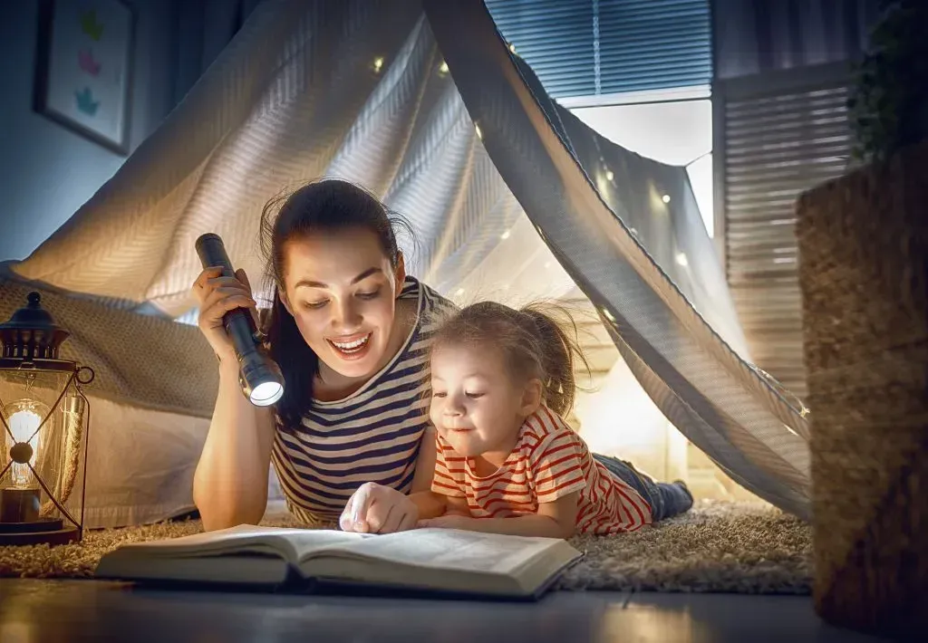 Mother and child reading a book in a blanket fort.