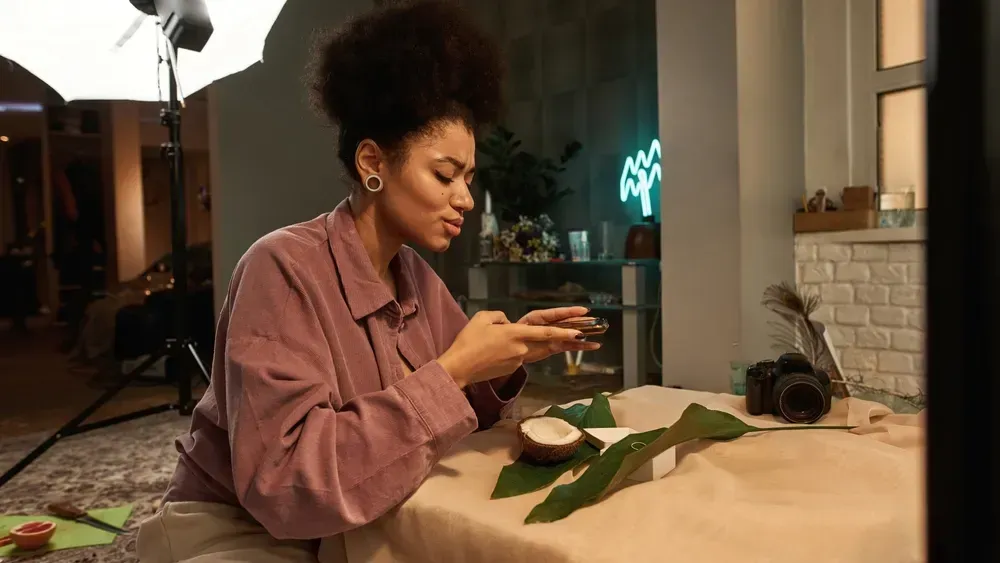 Side view of black female photographer taking photo of ring, big leaves, sliced coconut and open box on smartphone at home studio. 