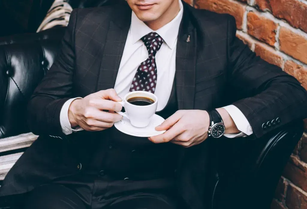 A man in a suit with a cup of coffee sits on a black leather sofa.