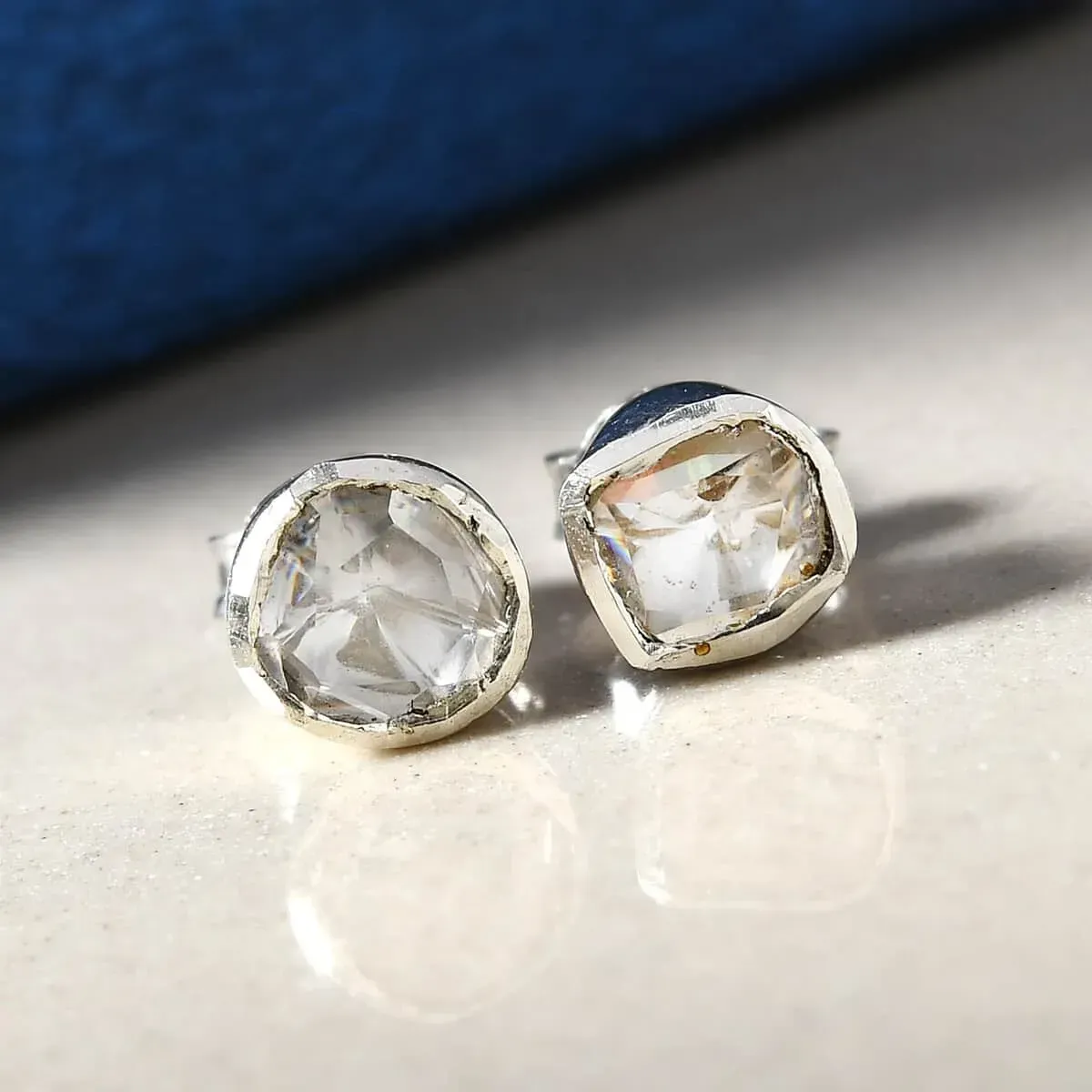 Polki Moissanite Solitaire Stud Earrings in Platinum Over Sterling Silver resting on glossy table with blue background.