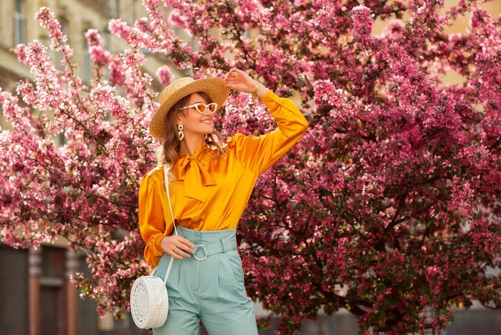 Happy smiling woman wearing trendy straw hat, yellow satin blouse, blue trousers, cat eye sunglasses, with wicker bag, posing in street near pink spring blossom trees.