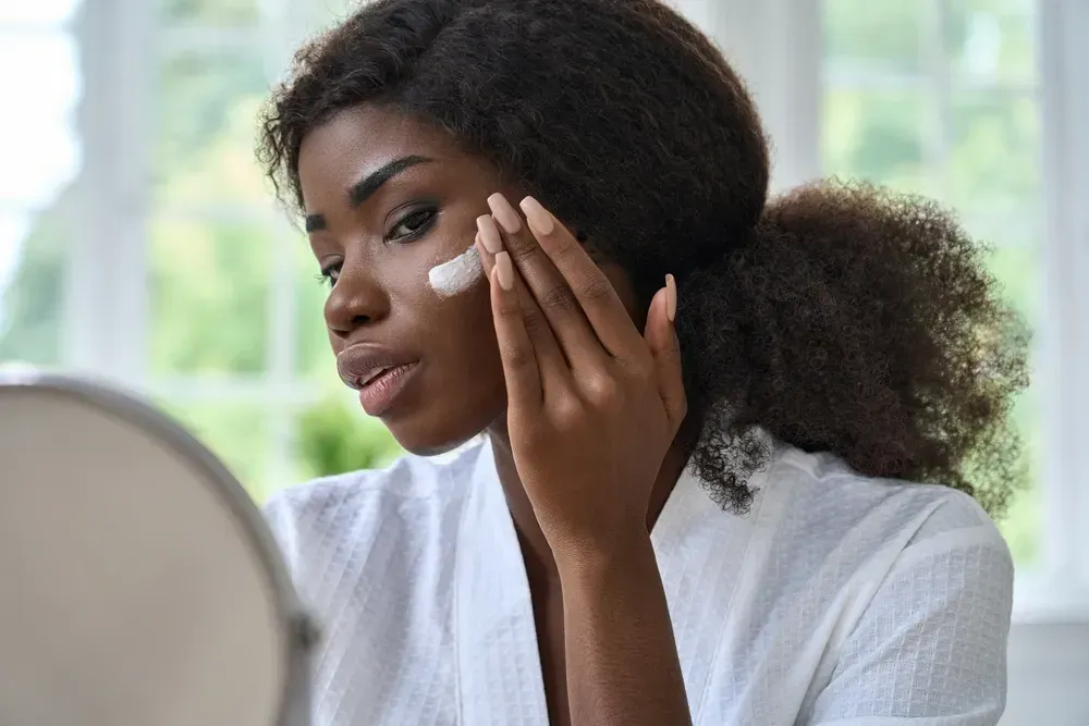 Attractive young black African woman model touching healthy flawless radiant smooth face skin care looking in bathroom mirror applying facial cream.