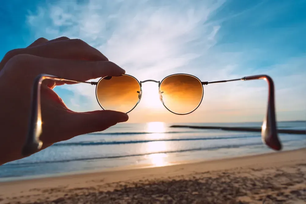 Hand holding stylish round sunglasses with brown lenses at sunset.
