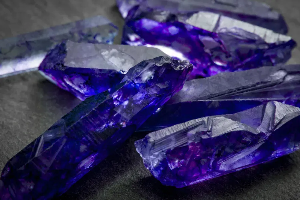 Raw tanzanite crystals, showing blue to violet color on charcoal gray background.