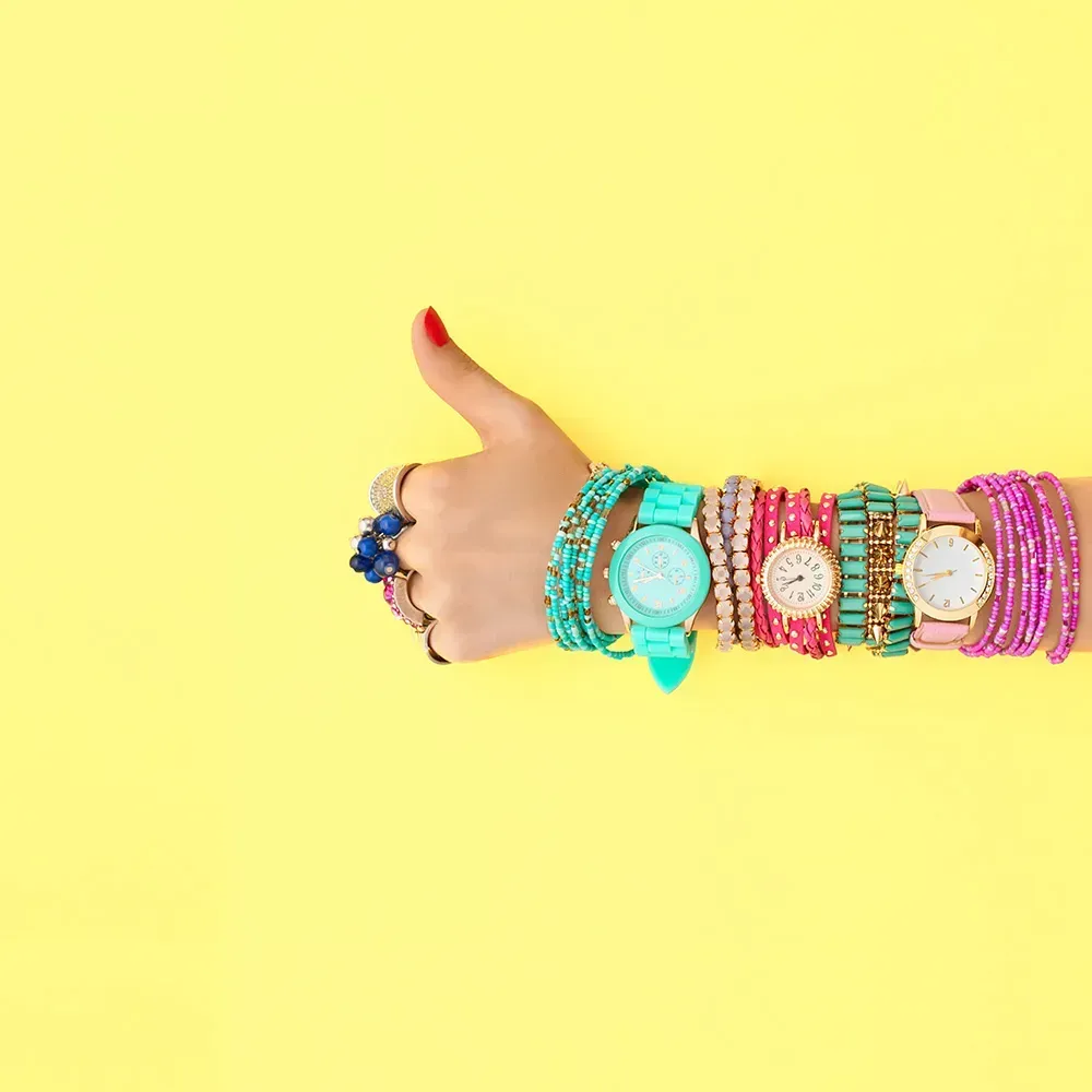 Closeup of woman wearing three colorful watches against yellow background giving a thumbs up.