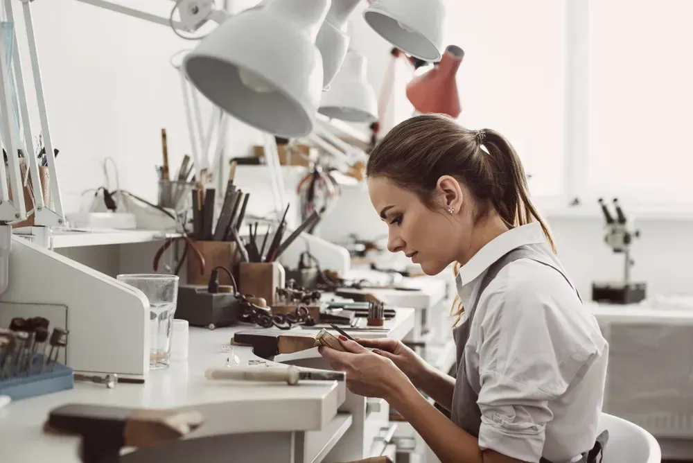 Working all day. Side view of young female jeweler sitting at her jewelry workshop and holding in hands jewelry tools for making accessories