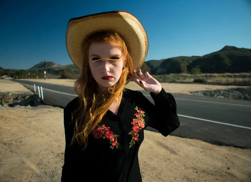 Young redhead woman in Western Gothic style, wearing an embroidered black button down shirt and cowboy hat standing in front of a lonely western highway.
